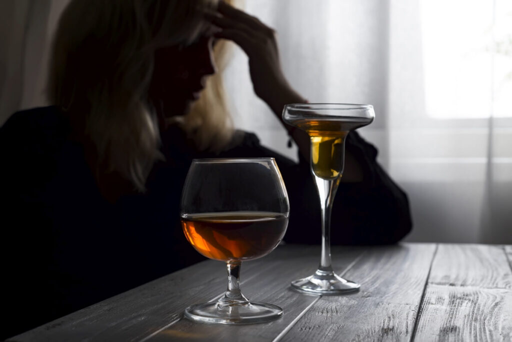 Kobieta z wyrazem smutku i frustracji siedząca przy stole, na którym znajdują się dwa kieliszki z alkoholem. Możliwe, że potrzebna jest terapia uzależnień.