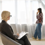 Terapeutka siedzi z notatnikiem naprzeciwko stojącej kobiety, rozmawiają o problemach związanych ze współuzależnieniem.
