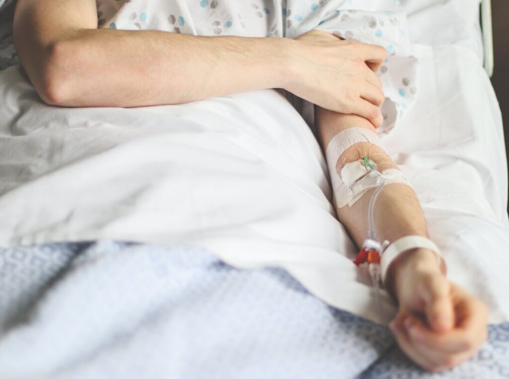 Osoba leży w szpitalnym łóżku z wenflonem w ramieniu, cierpiąc na bóle stawów.